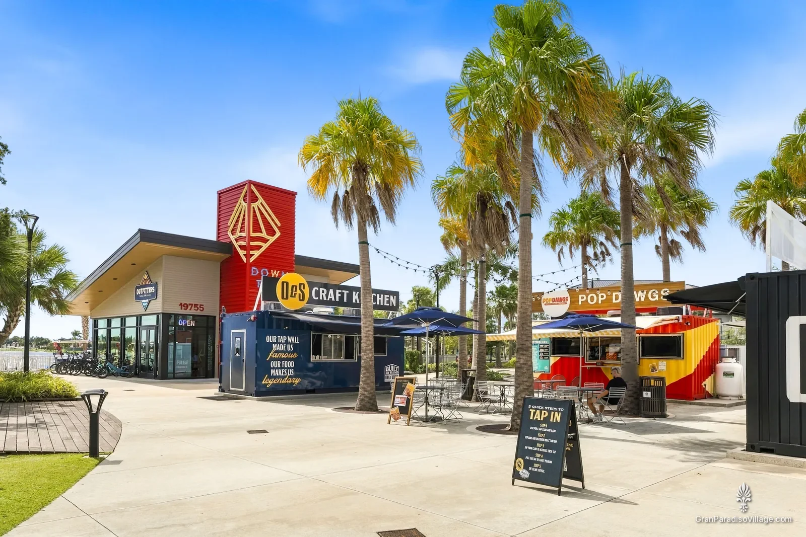 Food-truck court under tall palms in Downtown Wellen, with container kitchens, blue umbrellas and café tables