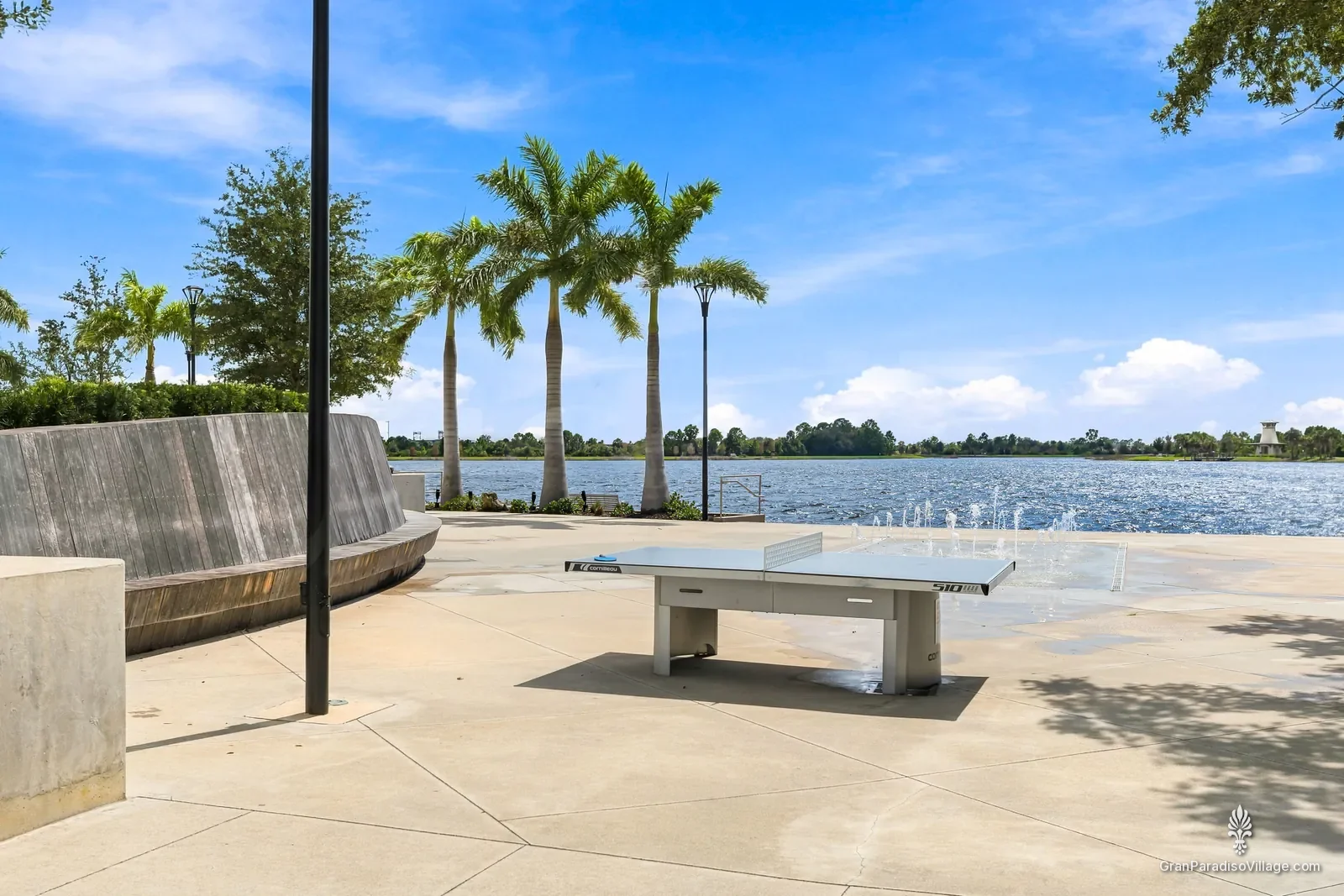 Outdoor ping-pong table and splash-pad fountains on the Downtown Wellen lakefront plaza, with royal palms and the Grand Lake