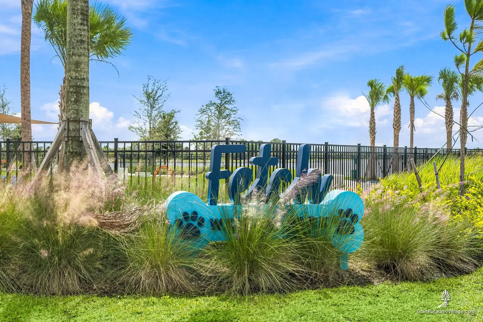 Blue Fetch Dog Park sign with paw prints among ornamental grasses, with the fenced dog park behind