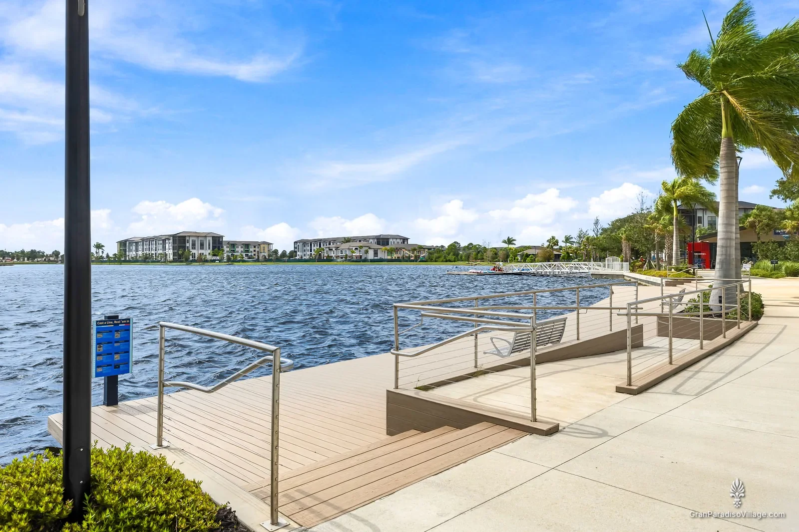 Wooden waterfront deck with stainless railings and steps down to the Grand Lake in Downtown Wellen, with palms and blue sky