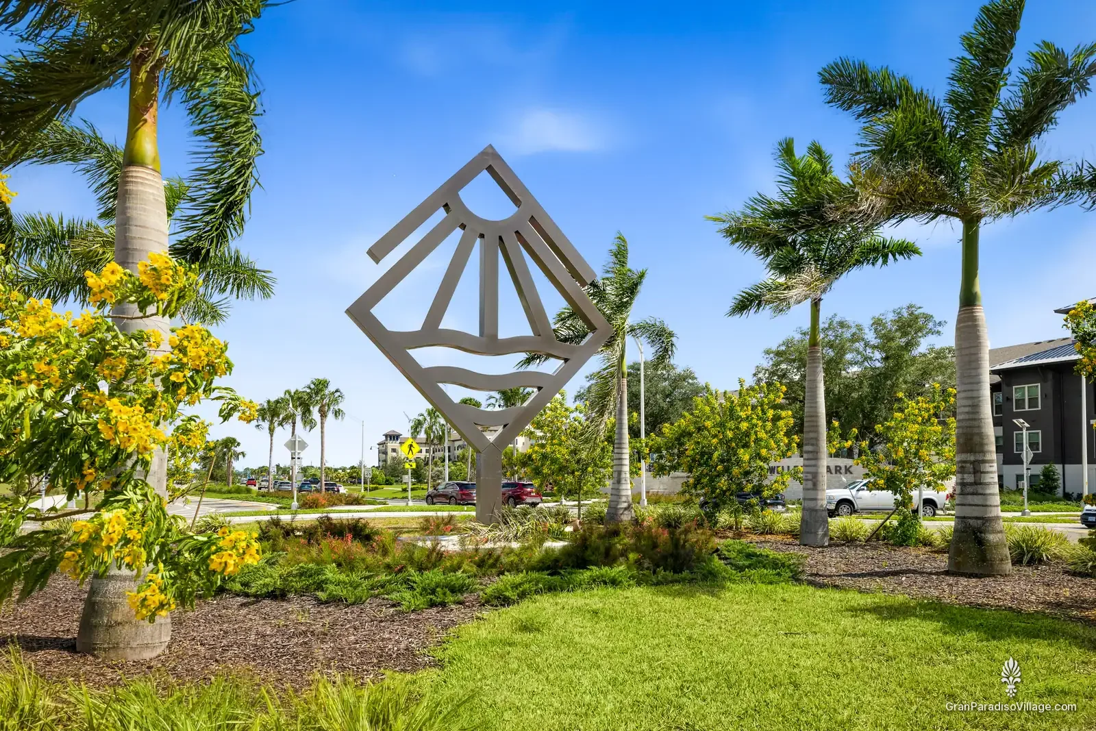 Wellen Park logo sculpture, a sun-and-waves diamond, in a landscaped bed with palms and yellow-flowering trees